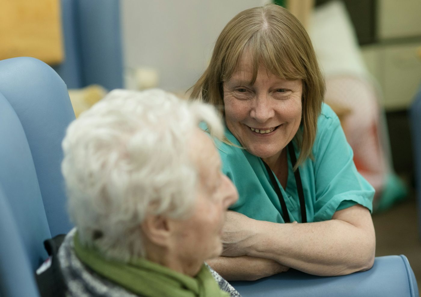 A caregiver in green scrubs leans in and smiles warmly while talking with an older woman seated in a care home