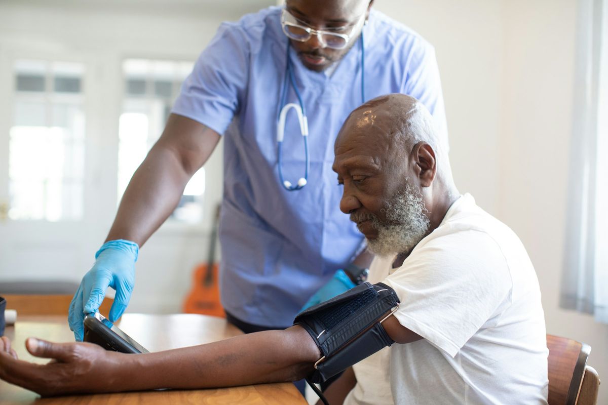 A nurse in blue scrubs and gloves checks the blood pressure of an older man seated at a table