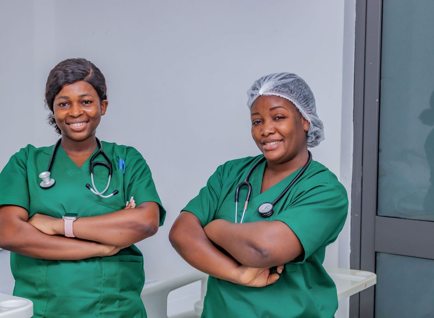 Two smiling nurses in green scrubs with stethoscopes stand with arms folded in a clinical room