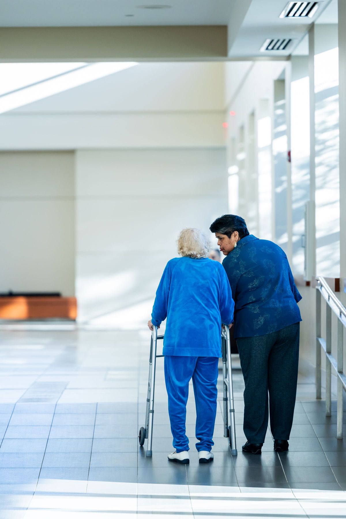 A caregiver walks alongside an older woman using a walker down a bright, sunlit facility hallway