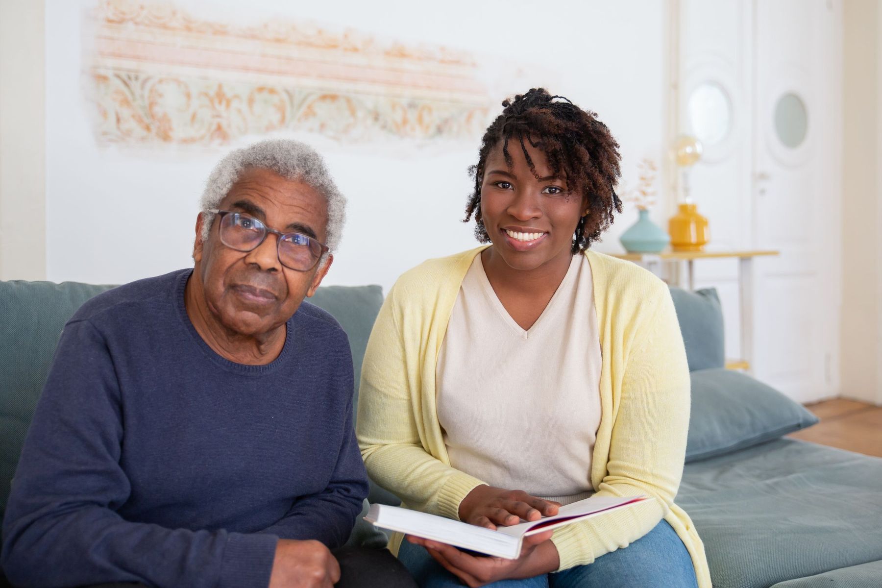 A smiling caregiver in a yellow cardigan sits beside an older man on a sofa, holding a book as they spend time together at home